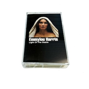 May include: A vintage cassette tape featuring Emmylou Harris' album "Light Of The Stable." The album cover displays a portrait of Harris with a white head covering against a black background. The cassette is in a clear plastic case.