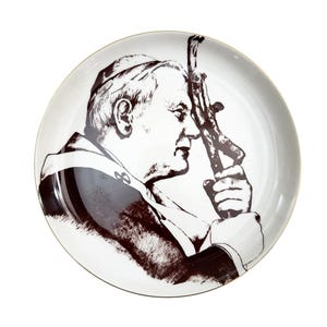 Puede incluir: Un plato de cerámica blanco con una ilustración marrón y negra de un hombre sosteniendo un crucifijo. El plato tiene un borde dorado y un acabado brillante. La ilustración es un retrato detallado.