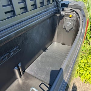 May include: A black truck bed storage compartment with a textured interior, a hinged lid, and a built-in electrical outlet labeled 115V AC MAX 400W. A metal tie-down loop is visible at the base of the storage area, which is integrated into the vehicle body.