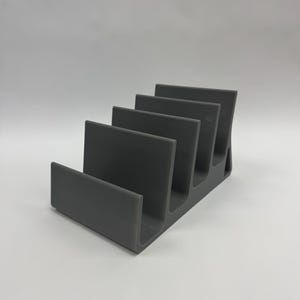 May include: A gray, stepped organizer with five slots. The organizer is made of a solid material and has a curved base. The slots are designed to hold items upright, such as mail, documents, or small books.