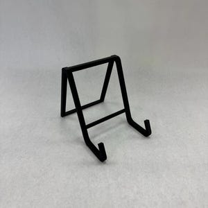 May include: A black metal display stand with a rectangular top and angled legs. The stand has a horizontal bar near the base and two curved arms to hold an item. The stand is designed to hold a small plate or picture.