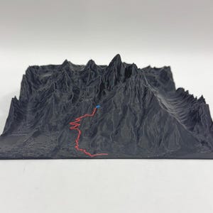 Puede incluir: Un mapa topográfico impreso en 3D en gris oscuro de una cordillera. Una línea roja traza un camino por la montaña, con un punto azul que marca un punto. El modelo está sobre una superficie blanca.
