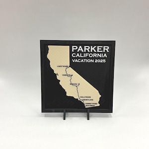 May include: A black framed wall art piece with a map of California in a light tan color. The text "PARKER CALIFORNIA VACATION 2025" is displayed above the map. The map highlights locations like Lake Tahoe, Yosemite, and Hollywood.