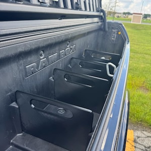 May include: A black plastic Ram Box truck bed storage compartment featuring multiple vertical dividers with integrated handle cutouts. The interior walls are molded with the embossed text RAM BOX, and a metal D-ring tie-down point is visible.