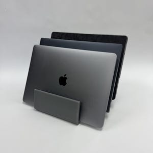 May include: Three gray laptops are displayed in a vertical stand. The front laptop has a black Apple logo. The stand is a matching gray color. The laptops are stacked, with the top one showing a dark pattern.