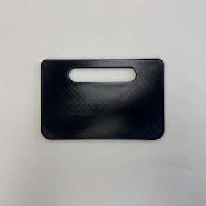 Op de afbeelding: Een rechthoekig zwart plastic object met afgeronde hoeken en een horizontale, langwerpige ovale uitsparing nabij de bovenrand. Het oppervlak heeft een subtiele diagonale kruisarcering, consistent met 3D-geprinte items, wat een vlak uiterlijk geeft.
