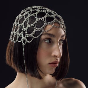 Könnte beinhalten: Ein silberfarbenes Kopfstück mit einem filigranen, offen gewebten Design. Eine passende Kette hängt vom Kopfstück herab und führt zu einer Halskette. Der Schmuck ist vor einem dunklen Hintergrund platziert.