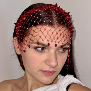 以下が含まれることがあります： 赤いビーズのヘッドピース、ネットデザイン。ヘッドピースはビーズの王冠と額を覆うネットが特徴です。ぶら下がっている赤いビーズがネットからぶら下がっています。エレガントなアクセサリーです。