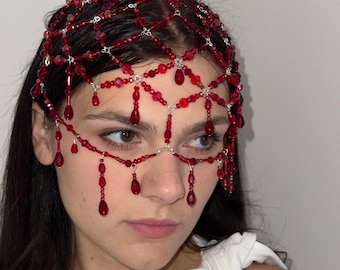 May include: A red beaded headpiece with a delicate, web-like design. The headpiece features small, round beads and larger, teardrop-shaped beads that dangle near the eyes. The intricate design is made with silver-coloured wire.