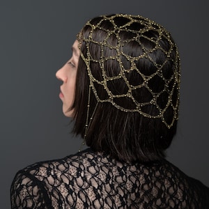 Puede incluir: Un tocado de cadena de metal en tono dorado con un diseño delicado en forma de red con pequeñas cuentas esféricas en cada unión, usado sobre cabello oscuro, combinado con una prenda de encaje negro de manga larga con patrón floral.