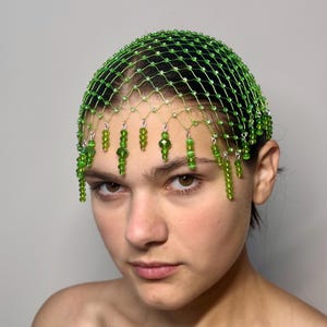 May include: A green beaded headpiece with a net design. The headpiece is adorned with dangling green beads. The accessory is designed to be worn on the head and is a unique fashion statement.