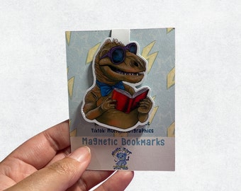 Op de afbeelding: Een magnetische bladwijzer met een cartoon-dinosaurus die een zonnebril en een blauwe strik draagt, en een rood boek vasthoudt. De bladwijzer heeft een blauwe en witte achtergrond met gele bliksemschichten. De tekst "Magnetic Bookmarks" staat op de bladwijzer gedrukt.