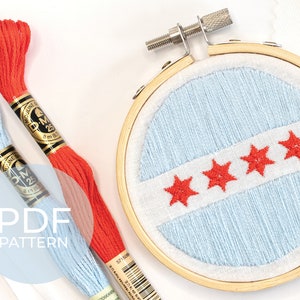 May include: A close-up of a finished embroidery hoop with a blue background and four red stars in a row. Two spools of embroidery floss are visible, one light blue and one orange.