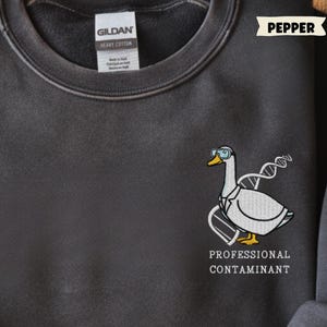 May include: Dark grey sweatshirt with a crew neck. The sweatshirt features an embroidered design of a goose wearing glasses and a lab coat, with the text "PROFESSIONAL CONTAMINANT" below. The sweatshirt is labeled "PEPPER" and "COMFORT COLORS".