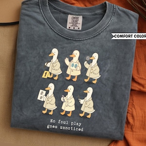 May include: Pepper-colored Comfort Colors t-shirt featuring a graphic of detective ducks. The ducks are depicted in beige trench coats, some holding magnifying glasses and evidence. The text "No foul play goes unnoticed" is printed below the ducks.
