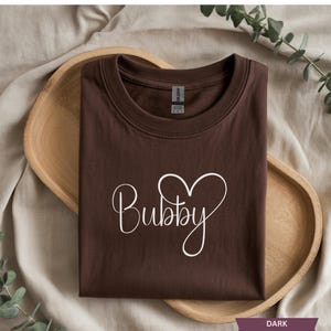 May include: Dark chocolate-colored t-shirt with the word "Bubby" in a white script font, with a heart-shaped design. The shirt is displayed on a wooden tray, with a neutral background and greenery.