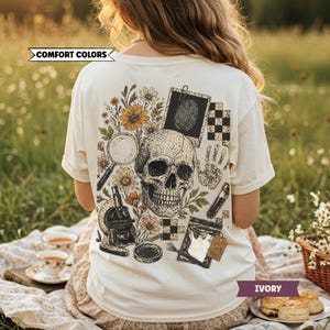 May include: Ivory-colored t-shirt with a graphic on the back featuring a skull, flowers, magnifying glass, microscope, and fingerprint. The text "COMFORT COLORS" is on the upper left, and "IVORY" is on the lower right.