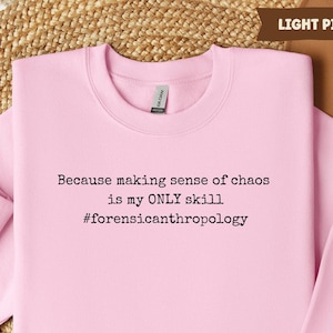May include: Light pink sweatshirt with the text "Because making sense of chaos is my ONLY skill #forensicanthropology" in black. The sweatshirt is a crew neck style.