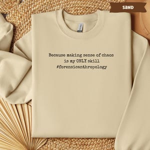 May include: Sand-colored sweatshirt with the text "Because making sense of chaos is my ONLY skill #forensicanthropology" in black. The sweatshirt has a crew neck and long sleeves. The word "SAND" is in a brown banner in the upper right corner.