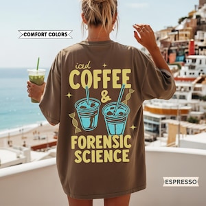 May include: Espresso-colored t-shirt with the text "iced COFFEE & FORENSIC SCIENCE" in yellow and blue. The shirt features two blue coffee cup illustrations with straws. The words "Comfort Colors" are also visible.