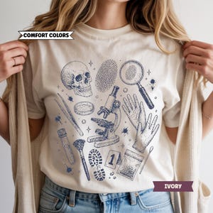 May include: Ivory-colored t-shirt featuring a forensic science design. The graphic includes a skull, fingerprints, magnifying glass, microscope, gloves, and other lab tools in a blue ink illustration. The text "COMFORT COLORS" and "IVORY" are also visible.