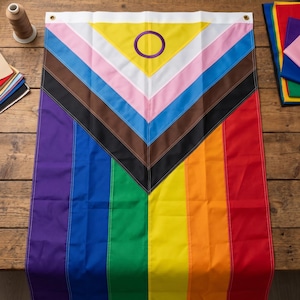 Könnte beinhalten: Eine rechteckige, intergeschlechtliche, inklusive Progress-Pride-Flagge mit vertikalen Streifen in Violett, Blau, Grün, Gelb, Orange und Rot. Ein Chevron-Muster überlagert den oberen Teil mit Weiß, Rosa, Hellblau, Braun, Schwarz und einem gelben Dreieck mit einem violetten Kreis.