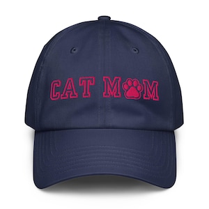 Puede incluir: Una gorra de béisbol azul marino con el texto bordado CAT MOM en una fuente de color rosa brillante. La letra O en la palabra MOM está reemplazada por un diseño de huella de pata bordado en rosa. La gorra tiene una corona estructurada.