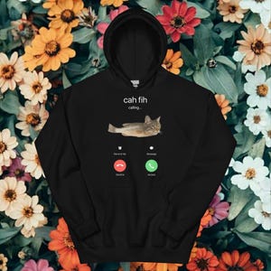 May include: Black hooded sweatshirt with a cat graphic and the text "cah fih calling...". The design mimics a phone call screen, showing "Decline" and "Accept" buttons. The background is a floral pattern.