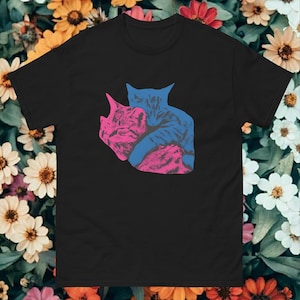 以下が含まれることがあります： 黒いTシャツ。ピンクと青の2匹の猫が抱き合っているグラフィック。猫はテクスチャ加工が施されています。柔らかい素材でできており、普段使いに適しています。