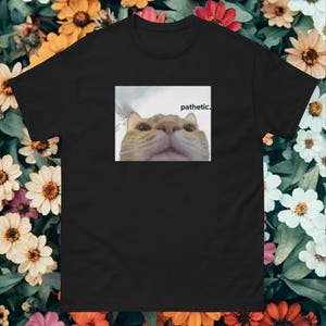 Könnte beinhalten: Schwarzes T-Shirt mit einem Meme eines Katzen-Gesichts und dem Wort "pathetic." darüber. Die Katze hat orange-weißes Fell. Das T-Shirt wird vor einem floralen Hintergrund mit verschiedenen Blumen gezeigt.
