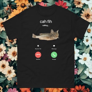 May include: Black t-shirt with a cat-fish hybrid graphic and the text "cah fih calling...". The design includes a phone interface with "Remind Me", "Message", "Decline", and "Accept" buttons. The cat-fish is brown and grey.