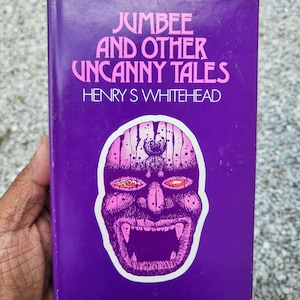 Op de afbeelding: Een vintage paperback boek getiteld "Jumbee and Other Uncanny Tales" van Henry S. Whitehead. De omslag is paars met roze letters en een roze illustratie van een monstergezicht met rode ogen.