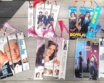 May include: A collection of handmade bookmarks featuring movie posters. Titles include "Father of the Bride," "My Best Friend's Wedding," and "Sleepless in Seattle." Each bookmark has a tassel attached at the top.