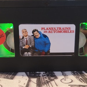 May include: A black VHS tape with a green light on each side. The front of the tape has a white label with the text "PLANES, TRAINS AND AUTOMOBILES" and a red sticker that says "CULT CLASSIC".
