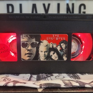 Puede incluir: Una lámpara de réplica de cinta VHS negra con una luz roja que brilla a través del frente y la parte posterior. La etiqueta frontal presenta el título de la película "The Lost Boys" y el texto "Cult Classic".