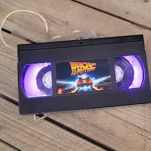 May include: A black VHS tape with a purple glow around the windows. The tape has a "Back to the Future" movie label with a picture of the DeLorean time machine.