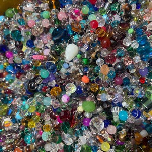 May include: A vibrant assortment of colorful beads in various shapes and sizes. The beads are a mix of clear, opaque, and iridescent, with colors including blue, green, pink, red, and yellow. Some beads are faceted, while others are smooth and round. A white teardrop-shaped bead is also present.