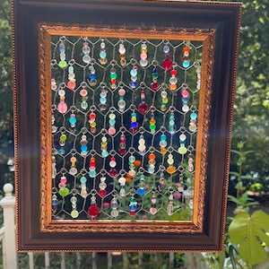 May include: A framed artwork featuring a wire mesh filled with colorful, dangling beads. The frame is dark brown with gold accents. The beads are various colors, including red, pink, blue, and green. The artwork is hanging outside.