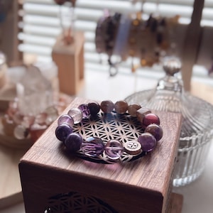 May include: A purple, brown, and clear crystal bracelet with a silver charm. The bracelet is on a wooden box with a geometric pattern.