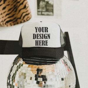 Może przedstawiać: Czarno-biała czapka typu trucker z napisem "YOUR DESIGN HERE" pogrubioną czcionką. Czapka spoczywa na lustrzanej kuli dyskotekowej, odbijającej światło. Tło obejmuje tkaninę w paski tygrysa i białą ścianę.