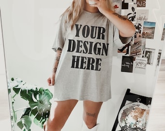 May include: A light gray oversized t-shirt with the text "YOUR DESIGN HERE" in black block letters. The person is wearing a black baseball cap and white knee-high boots. A disco ball and black chair are visible in the mirror's reflection.