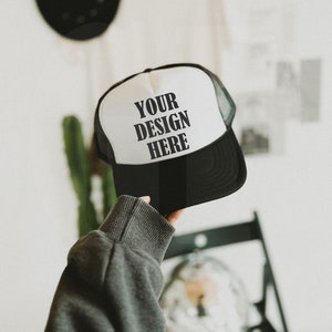 Może przedstawiać: Czarno-biała czapka typu trucker z napisem "YOUR DESIGN HERE" wydrukowanym z przodu. Czapka ma siatkowy tył i zakrzywiony daszek. Czapka jest trzymana przez osobę.