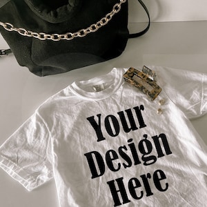 May include: White t-shirt with the text "Your Design Here" in black. A black bag with a gold chain strap is in the background. A leopard print hair clip and two gold earrings are on the shirt.