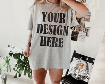 May include: A light gray t-shirt with the text "YOUR DESIGN HERE" in black block letters. The person in the image is wearing a black baseball cap and white boots. A disco ball sits on a black chair.
