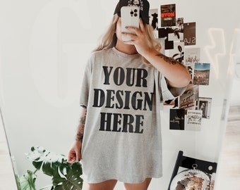 May include: A gray t-shirt with the text "YOUR DESIGN HERE" in black block letters. The person in the image is wearing a black baseball cap and white boots. A mirror reflects the scene, with a disco ball and posters visible.