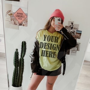 Può includere: Una maglietta verde neon con la scritta "YOUR DESIGN HERE" è indossata con una giacca di pelle nera, un berretto rosa e stivali da cowboy bianchi e neri. Un cactus in un vaso bianco è visibile nello specchio.