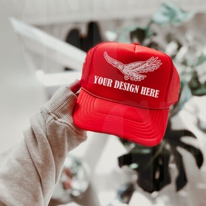 Może przedstawiać: Czerwona czapka typu trucker z białą grafiką orła i napisem "YOUR DESIGN HERE". Czapka ma siatkową tylną część i zakrzywiony daszek. Czapka jest trzymana przez osobę.