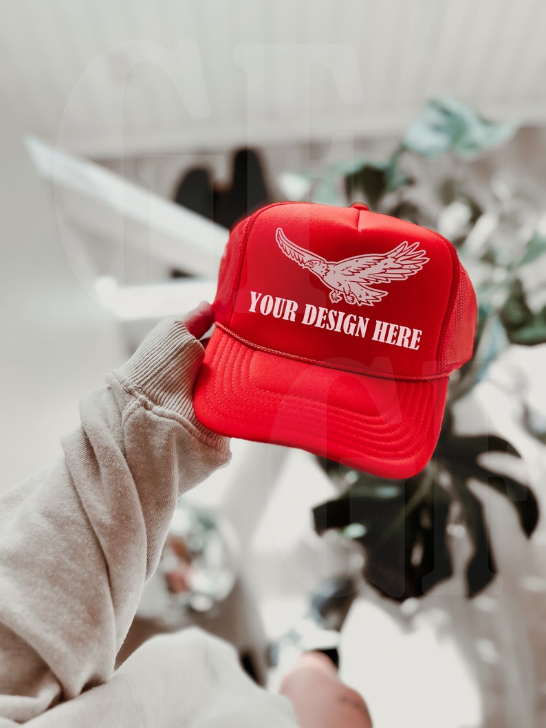 Może przedstawiać: Czerwona czapka typu trucker z białą grafiką orła i napisem "YOUR DESIGN HERE". Czapka ma siatkową tylną część i zakrzywiony daszek. Czapka jest trzymana przez osobę.
