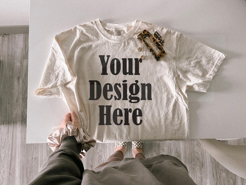 May include: Cream-colored t-shirt with the text "Your Design Here" in bold black letters. A tortoiseshell hair clip and small gold earrings rest on the shirt. The shirt is on a white surface.