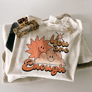 Puede incluir: Una camiseta color crema con el texto "You are Enough" en naranja y marr&oacute;n, con un sol y una nube sonrientes. Una pinza para el pelo carey y pendientes dorados descansan sobre un su&eacute;ter doblado.